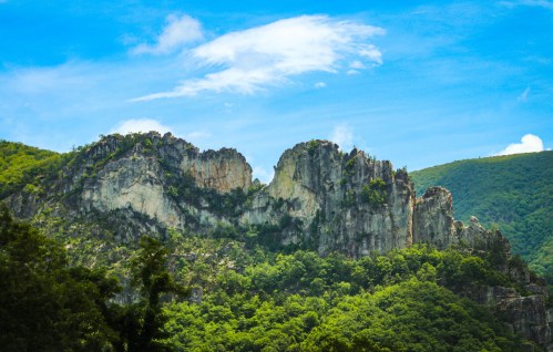 Majestic rocky peaks rise above lush green hills under a bright blue sky with scattered clouds.