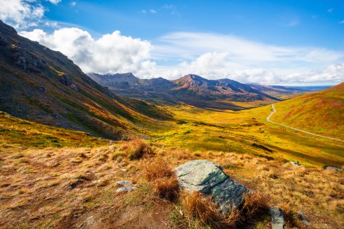 A scenic view of a valley surrounded by mountains, featuring vibrant autumn foliage under a blue sky with clouds.