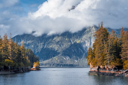 A serene landscape featuring a calm waterway surrounded by mountains and trees under a cloudy sky.