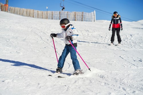 A child skis down a snowy slope, while an adult watches from behind on a sunny day.