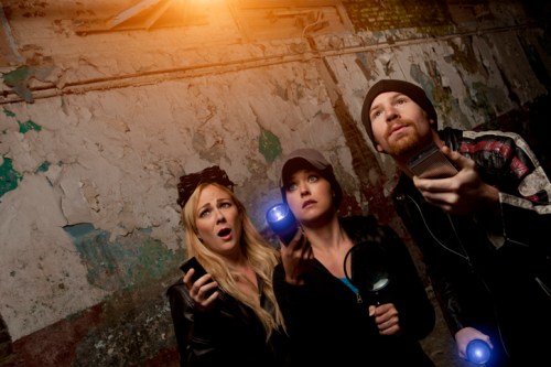 Three people in a dimly lit, abandoned space, looking up in surprise while holding flashlights and phones.