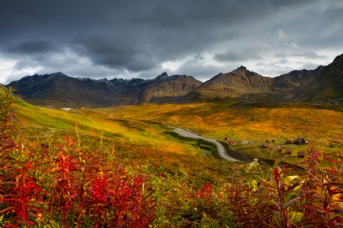 A scenic landscape featuring mountains, a winding road, and vibrant autumn foliage under a dramatic sky.