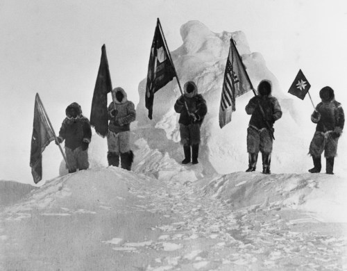 Five figures in winter gear stand on a snowy peak, holding various flags against a stark, icy landscape.