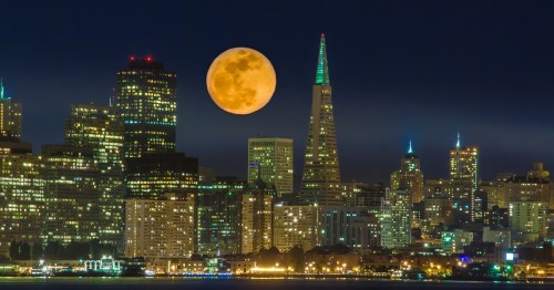 A glowing full moon rises over a city skyline, featuring tall buildings and a prominent spire, illuminated at night.