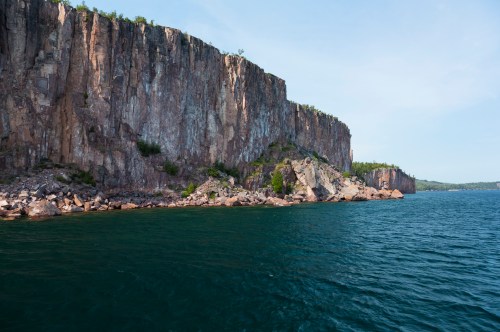 A rugged cliff rises above a calm blue lake, surrounded by greenery under a clear sky.