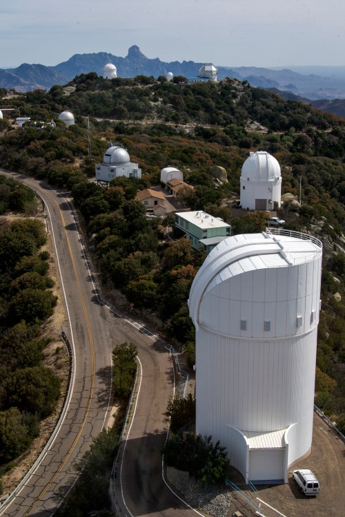 Aerial view of observatories on a hillside, with a winding road and mountains in the background.