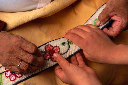 Two hands, one elderly and one young, work together on a colorful beaded textile with floral designs.