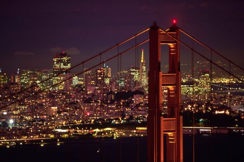 Golden Gate Bridge illuminated at night, with the San Francisco skyline sparkling in the background.