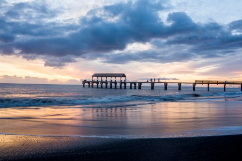 A serene beach scene at sunset, featuring a wooden pier extending into calm waters under a cloudy sky.