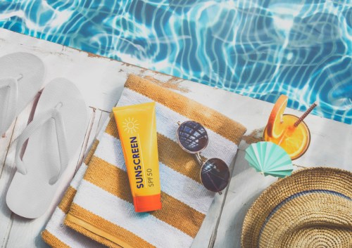 A poolside scene featuring sunscreen, sunglasses, a drink, flip-flops, and a towel on a sunny day.