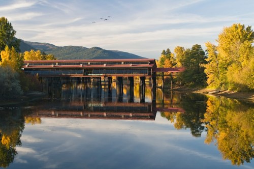 A modern building on stilts reflects in a calm river, surrounded by autumn trees and a clear blue sky.