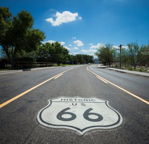 A clear view of a road with a "Historic US 66" sign painted on it, surrounded by trees and a blue sky.