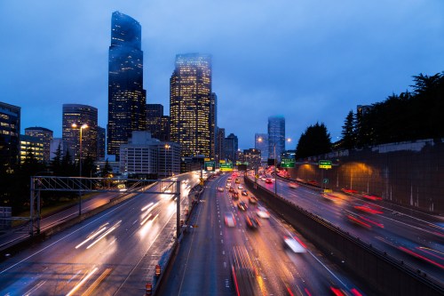 City skyline at dusk with tall buildings, a busy highway, and light trails from moving cars under a cloudy sky.