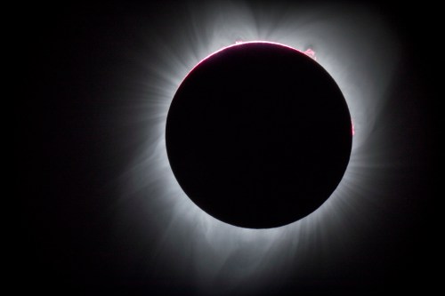 A solar eclipse with a darkened sun surrounded by a glowing corona and faint solar flares.