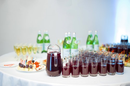 A table set with glasses of dark beverage, sparkling water bottles, and a platter of assorted snacks.