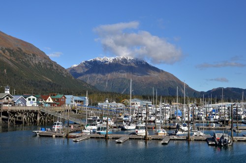 A scenic harbor with boats docked, surrounded by colorful buildings and mountains under a clear blue sky.