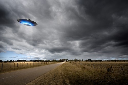 A UFO hovers above a rural road under a dark, cloudy sky, with fields on either side.