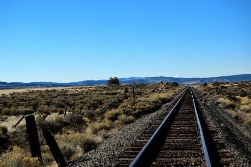 A view of railroad tracks stretching into the distance, surrounded by sparse vegetation and mountains under a clear blue sky.