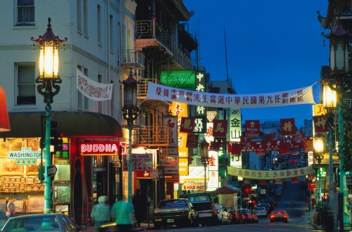 A vibrant Chinatown street at dusk, featuring colorful signs, lanterns, and festive banners.
