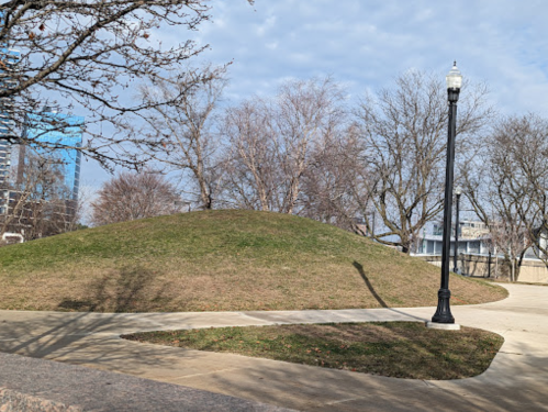 A grassy hill beside a paved path, with bare trees and a streetlamp in a park setting under a cloudy sky.