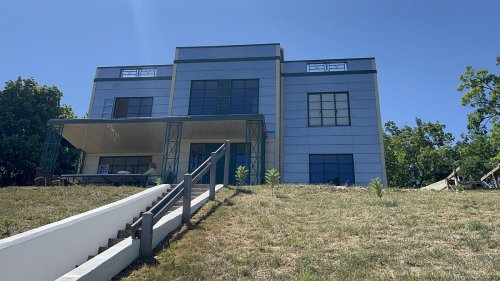 Modern two-story house on a hillside, featuring large windows and a grassy lawn under a clear blue sky.