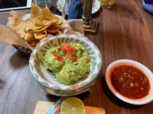 A bowl of guacamole topped with tomatoes, served with tortilla chips and a side of salsa on a wooden table.