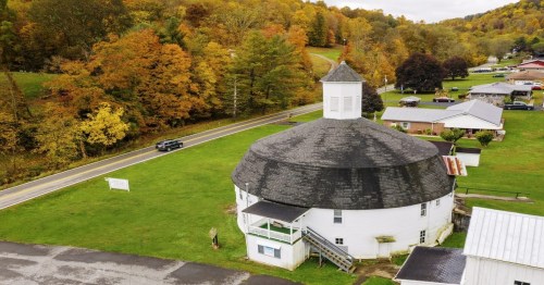 Aerial view of a round barn surrounded by colorful autumn trees and a road in a rural setting.