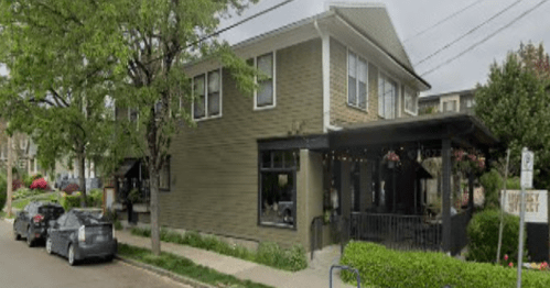 A two-story green house on a quiet street, with parked cars and trees lining the sidewalk.