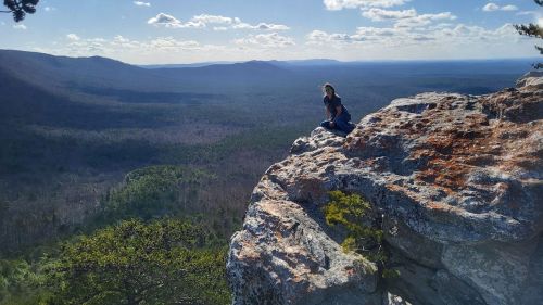 A person sits on a rocky ledge overlooking a vast forested valley under a blue sky with scattered clouds.