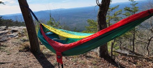 A colorful hammock hangs between two trees, overlooking a scenic mountain landscape on a sunny day.