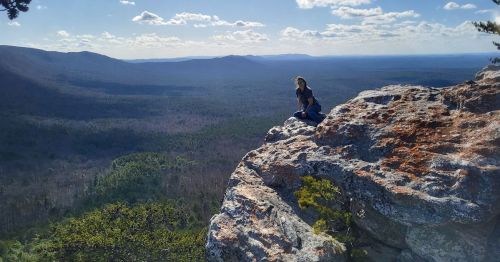 A person sits on a rocky ledge overlooking a vast forested landscape under a blue sky with scattered clouds.