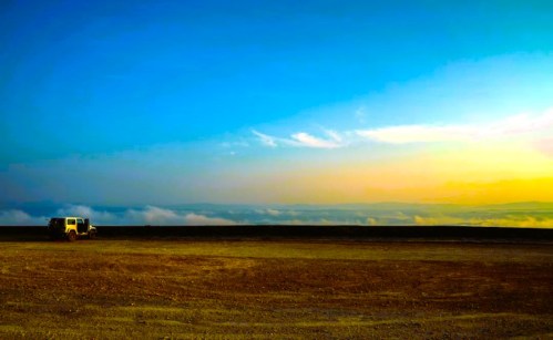 A lone vehicle parked on a vast, open landscape under a colorful sky at sunset, with clouds in the distance.