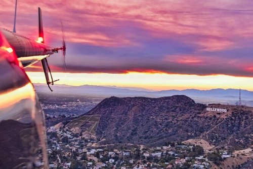 A helicopter view of the Hollywood sign at sunset, with vibrant clouds and mountains in the background.