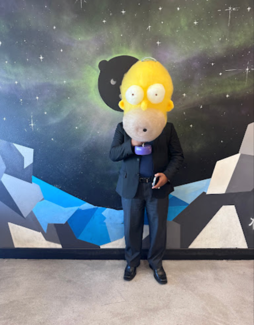 A person in a large Homer Simpson mask stands in front of a colorful mural, holding a microphone.