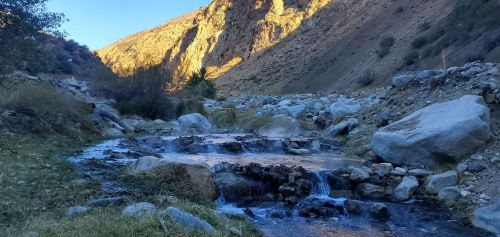 A serene mountain stream flows through rocky terrain, surrounded by steep hills and sunlight illuminating the landscape.