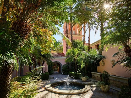 Lush courtyard with palm trees, a fountain, and a tower in the background, bathed in sunlight.