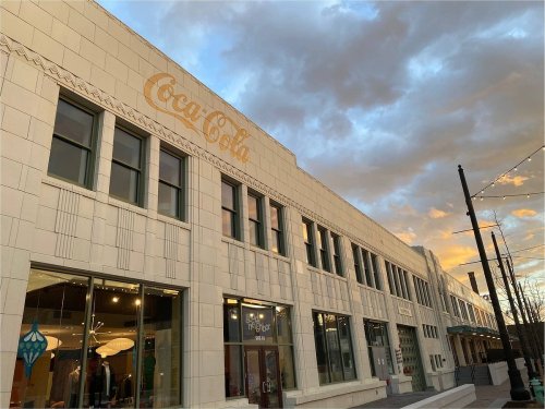 Coca-Cola building with large logo, featuring a modern facade and cloudy sunset sky in the background.