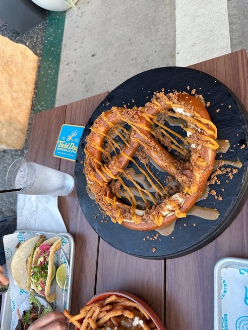 A large pretzel drizzled with mustard and topped with crumbled meat, served on a black platter with sides of tacos and fries.