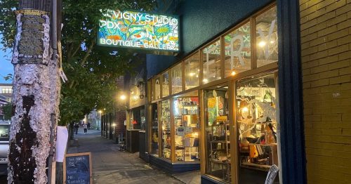 A boutique gallery with colorful signage, showcasing art and crafts, on a city street at dusk.