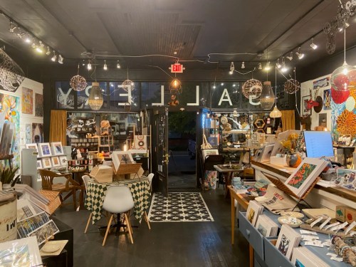 A cozy art gallery interior filled with colorful artwork, crafts, and decorative items, illuminated by warm lighting.