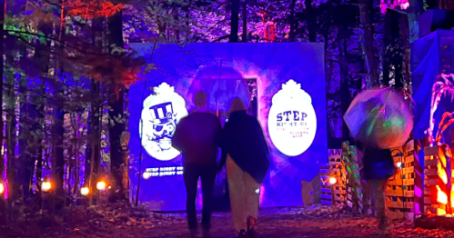 A couple walks towards a brightly lit entrance in a forest, surrounded by colorful lights and decorations.