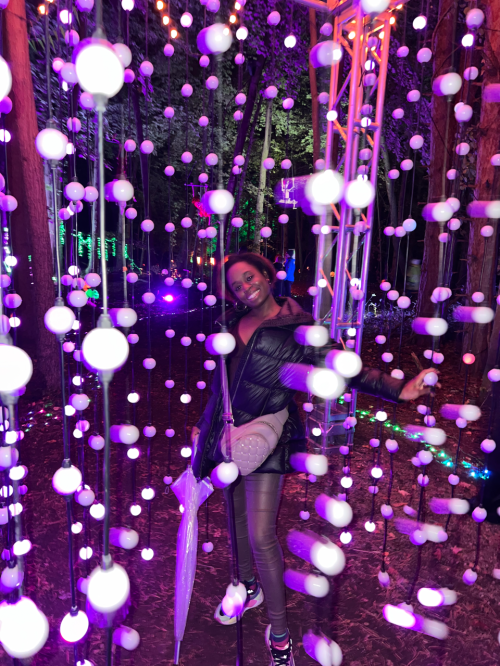 A person poses joyfully among colorful lights and hanging decorations in a festive outdoor setting.