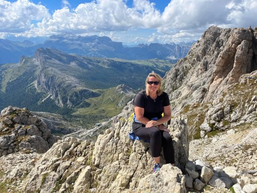 A woman sits on a rocky outcrop, smiling against a backdrop of mountains and blue skies.