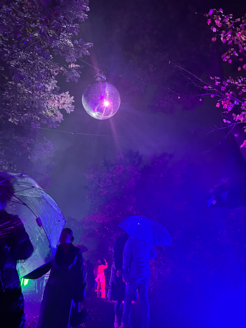 A crowd with umbrellas stands under a large disco ball, surrounded by colorful lights and misty night atmosphere.