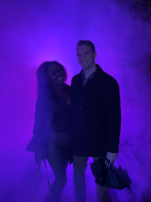 A man and woman stand together in a purple fog, smiling against a vibrant, colorful background.