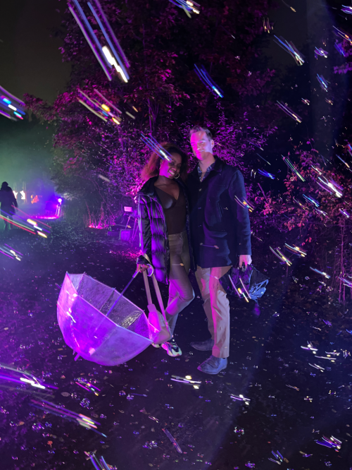 A couple stands under colorful lights, holding a clear umbrella, surrounded by a dreamy, vibrant atmosphere.