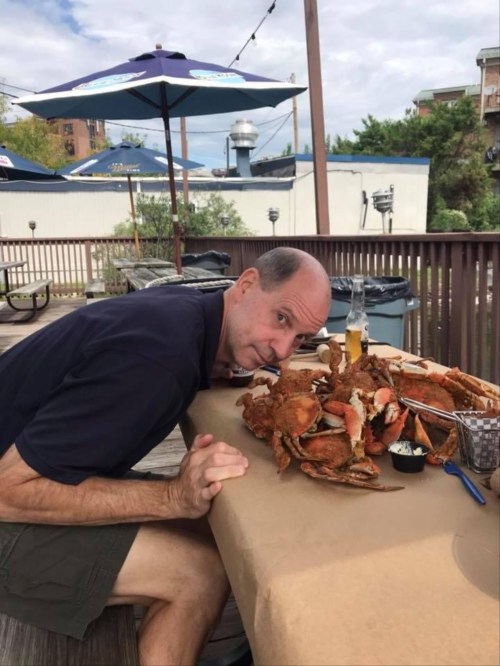 A man leans over a large pile of crabs on a table, with a drink nearby and a casual outdoor setting in the background.