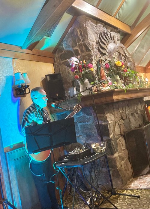 A musician performs with a guitar in front of a stone fireplace adorned with flowers and decorative masks.
