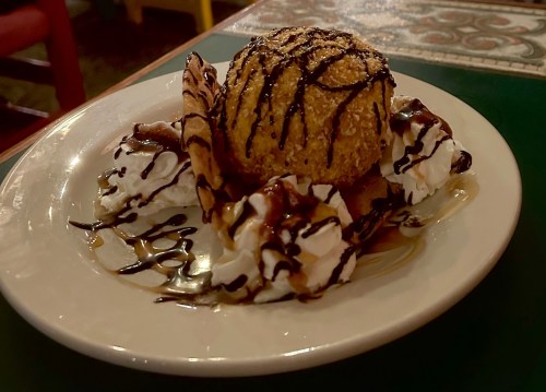 A dessert plate featuring a large fried ice cream ball, drizzled with chocolate, surrounded by whipped cream and a cookie.