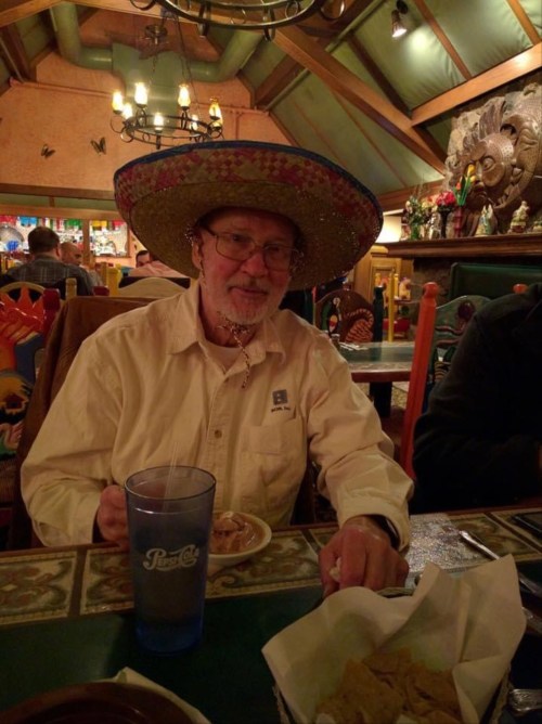 An older man wearing a colorful sombrero sits at a table with food and a drink in a festive restaurant setting.
